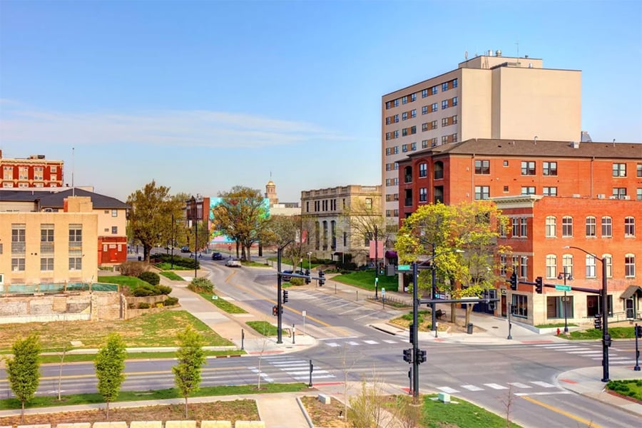 A quiet city intersection with brick and beige buildings, trees, crosswalks, and traffic lights on a clear, sunny day.