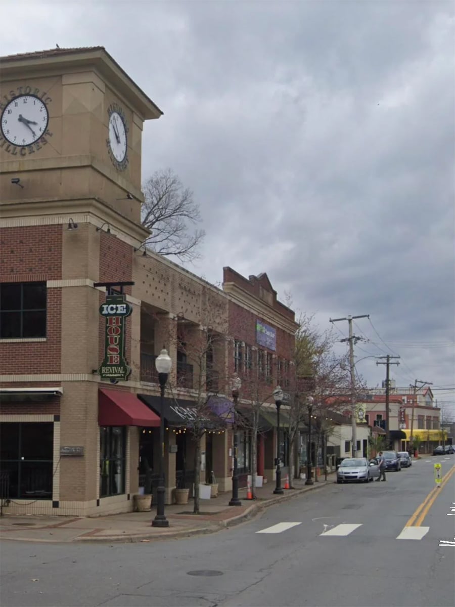 A brick building with a clock tower and “ICE HOUSE” rental sign stands by a crosswalk on a cloudy day. Trees without leaves line the sidewalk along a quiet street.