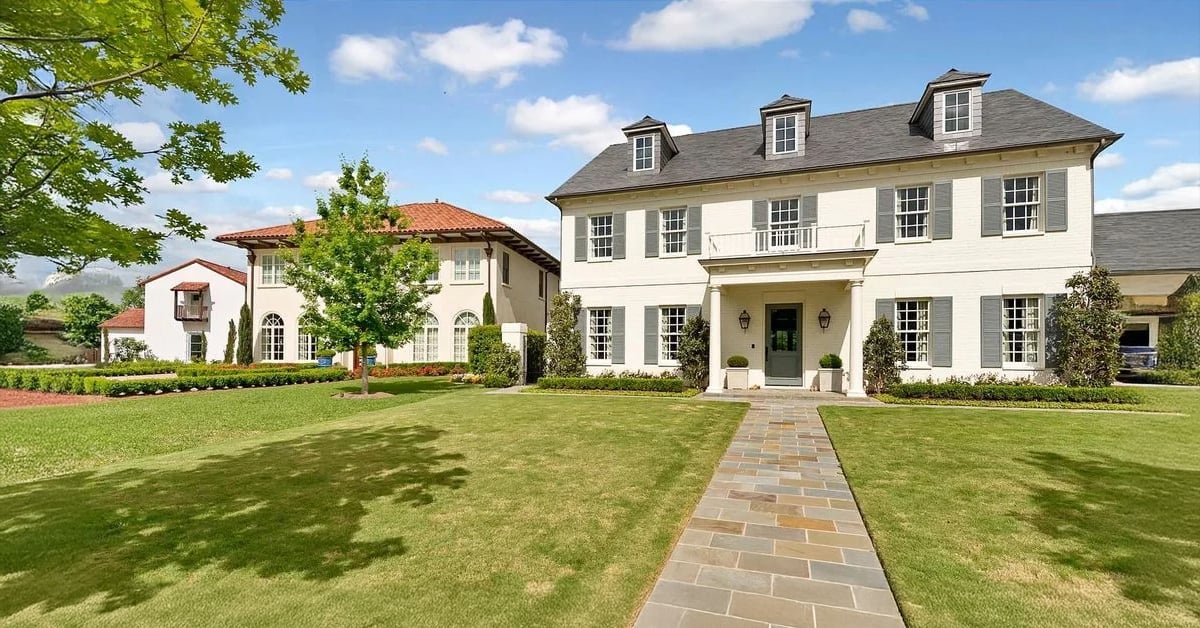 Two large, elegant houses with manicured lawns and stone walkway under a blue sky with clouds; trees and bushes decorate the front yards.