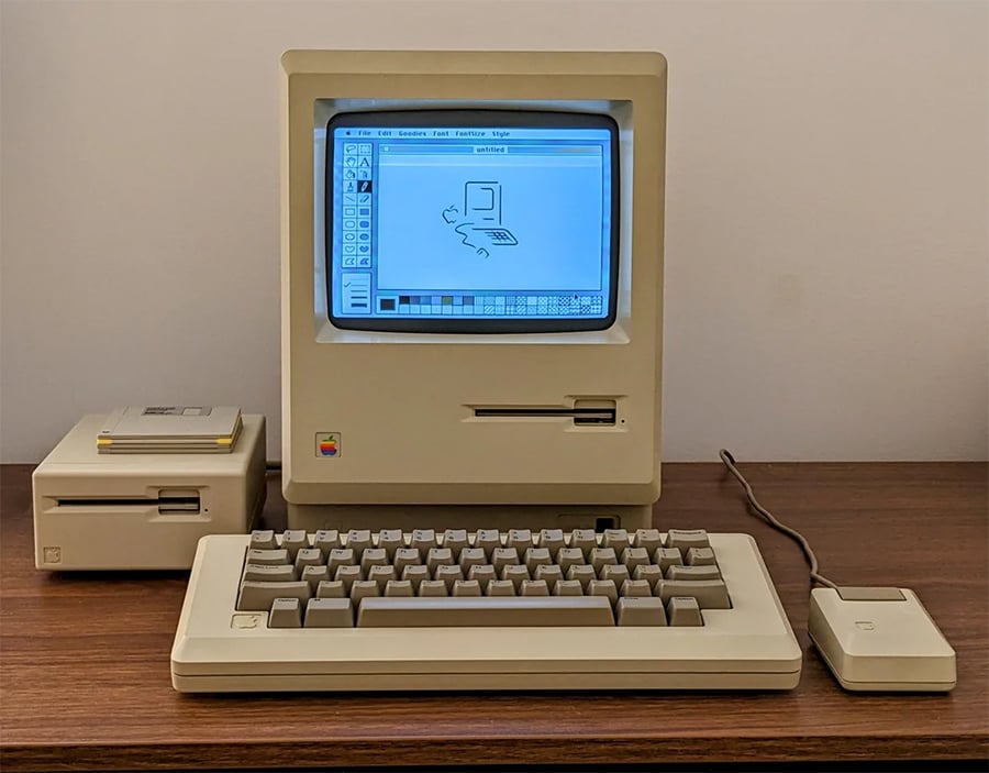A vintage Macintosh computer with monitor, keyboard, mouse, and external floppy disk drive sits on a wooden desk. The screen displays an early graphical interface.