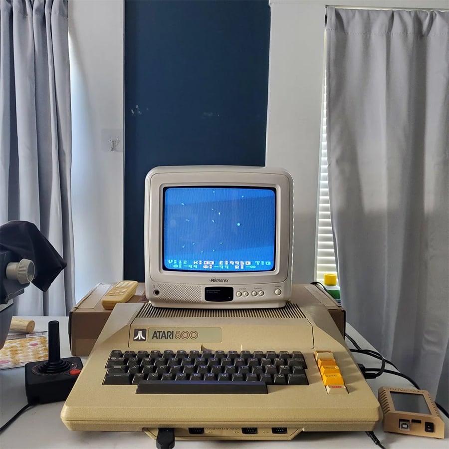 An Atari 800 computer with a joystick and monitor sits on a white desk. The screen displays a blue background with star-like dots. Curtains and a window are in the background.