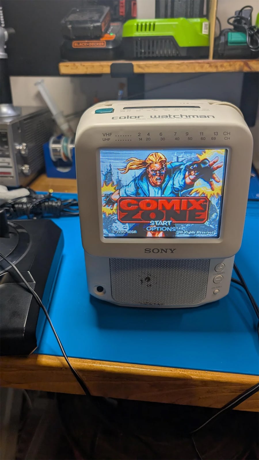A small Sony TV on a blue mat displays a colorful video game screen featuring two characters and the word “COMIX” in bold letters. Electronic equipment and cables surround the TV on a wooden table.