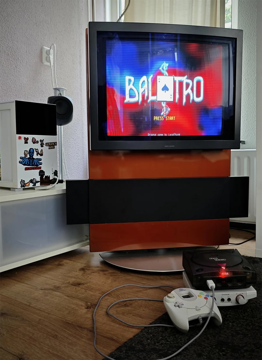 A Sega Dreamcast with controller is connected to a TV displaying the game “Balatro” title screen. The setup sits on a wooden floor next to a white shelf decorated with gaming stickers.