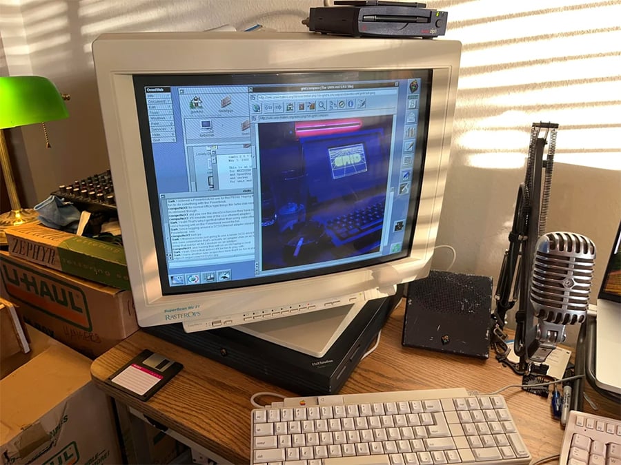 A vintage computer with a CRT monitor displays a photo editor. A beige keyboard, floppy disks, microphone, lamp, U-Haul boxes, and other electronics are on the wooden desk. Sunlight casts shadows on the wall.