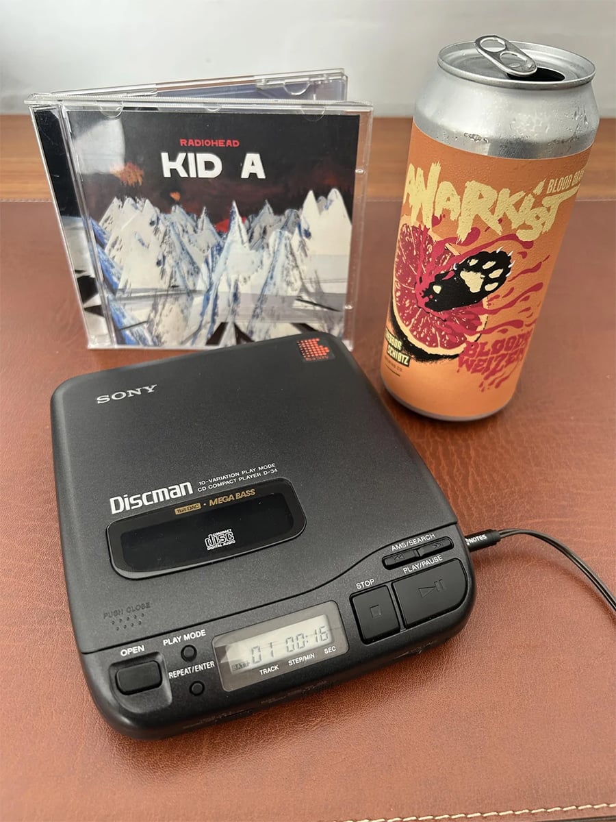 A Sony Discman playing a CD, with the Kid A album by Radiohead in the background and a can of beer with a vibrant label featuring a pomegranate design, all on a brown leather surface.