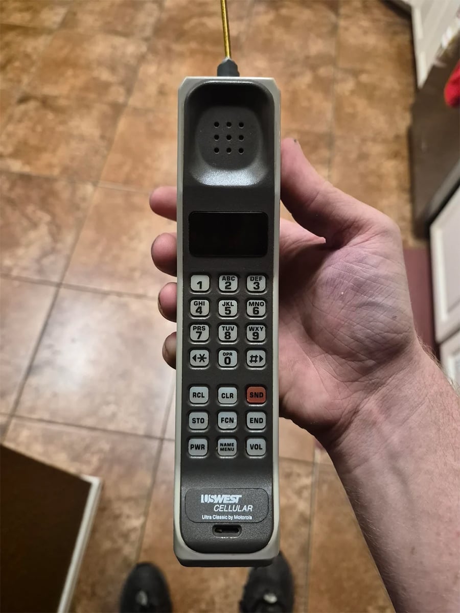 A hand holds a vintage US West Cellular mobile phone with a physical keypad and antenna, shown over a brown tiled floor.
