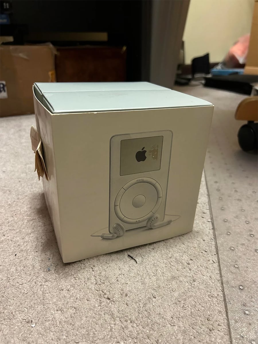 A white box featuring an illustration of an early Apple iPod with earbuds sits on a carpeted floor. Some packages and office furniture are visible in the background.