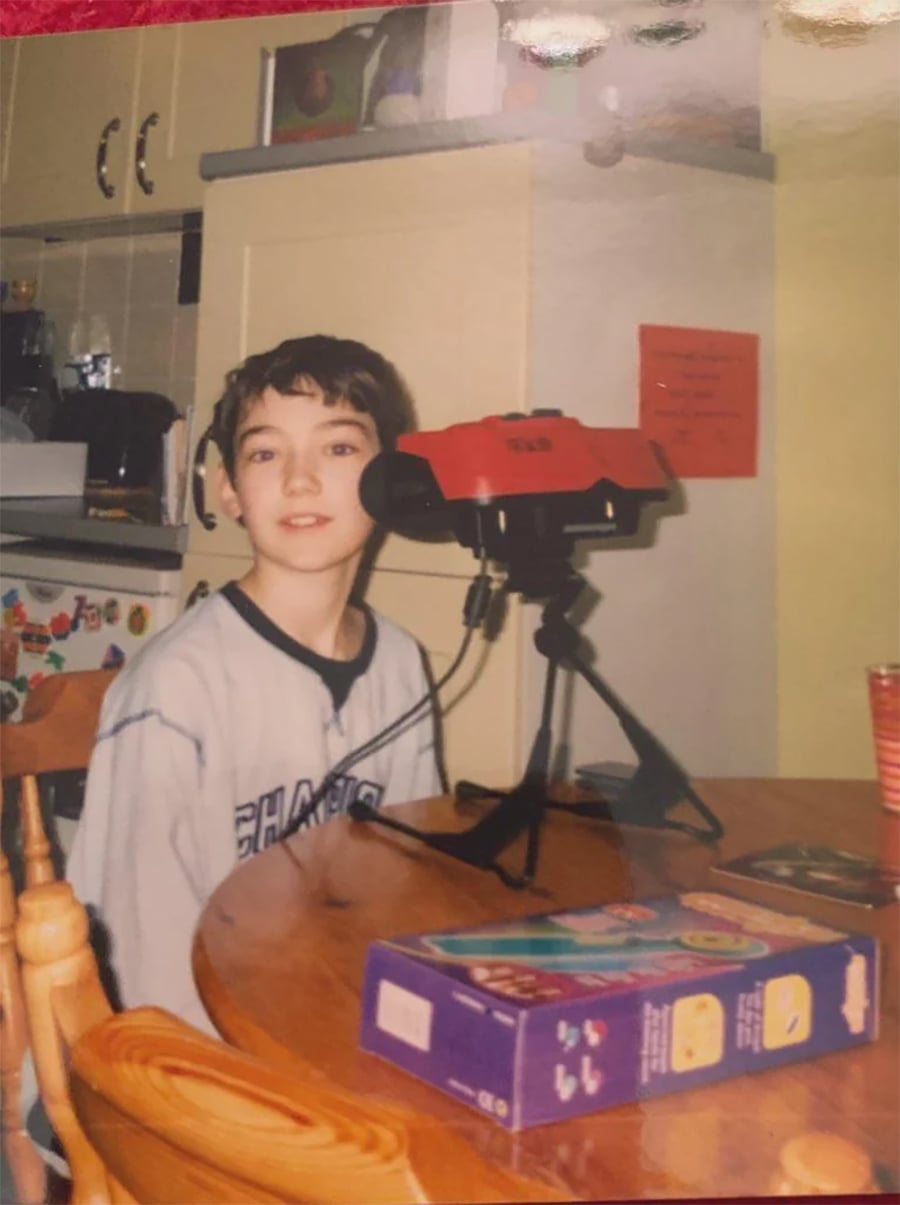 A boy sits at a kitchen table with a Nintendo Virtual Boy console in front of him. There are game boxes and a red cup on the table, and a fridge with magnets is visible in the background.
