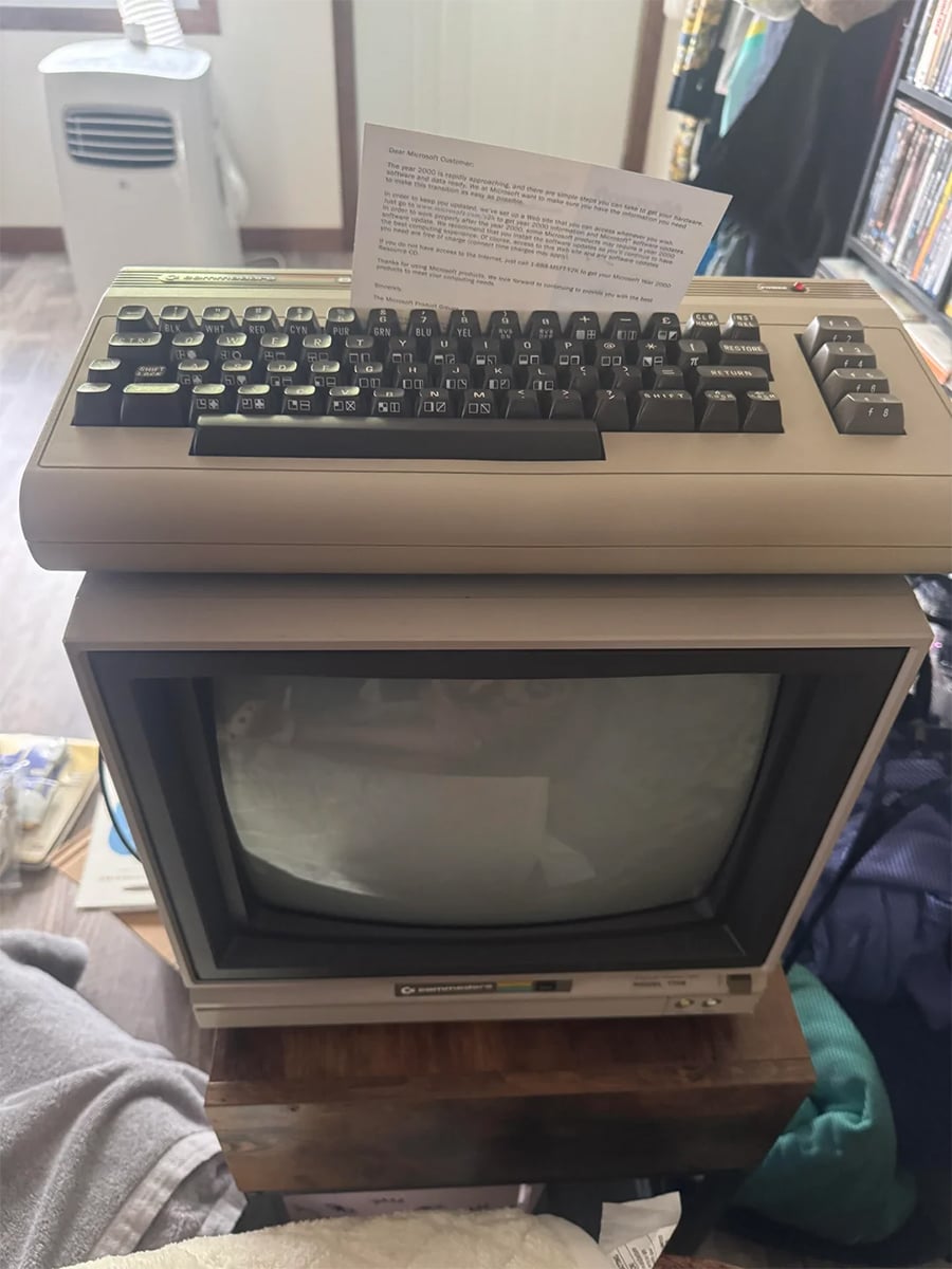 A vintage Commodore 64 computer sits on top of an old CRT monitor, with a printed note placed on the keyboard. The setup is indoors, surrounded by various household items.