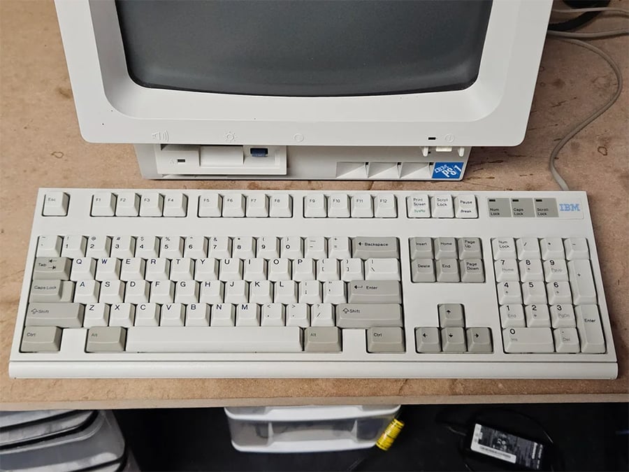 A vintage IBM keyboard with beige and gray keys is placed in front of an old IBM computer monitor and disk drive on a wooden desk. The scene has a retro, nostalgic tech feel.