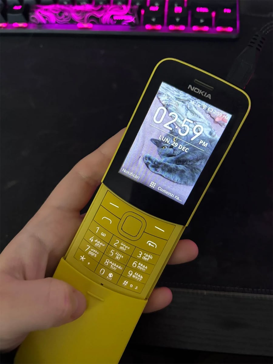 A hand holds a yellow Nokia slider phone displaying the time 02:59 PM on December 30, with a cat photo as the wallpaper. A keyboard with pink lighting is visible in the background.