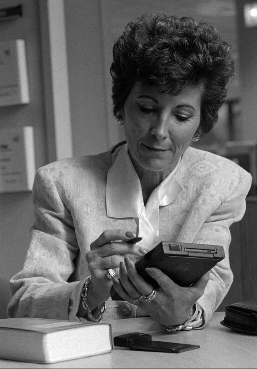 A woman in a patterned blazer uses a stylus on a handheld electronic device at a desk, with a closed book and another small object nearby.