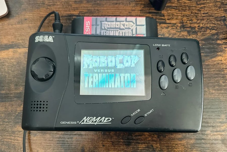 A Sega Genesis Nomad handheld console displaying the title screen for “RoboCop Versus The Terminator,” with the game cartridge inserted. The console rests on a wooden surface.
