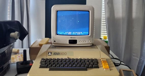 An Atari 800 computer with a joystick and a monitor displaying a blue screen sits on a desk, surrounded by curtains and other electronic equipment.