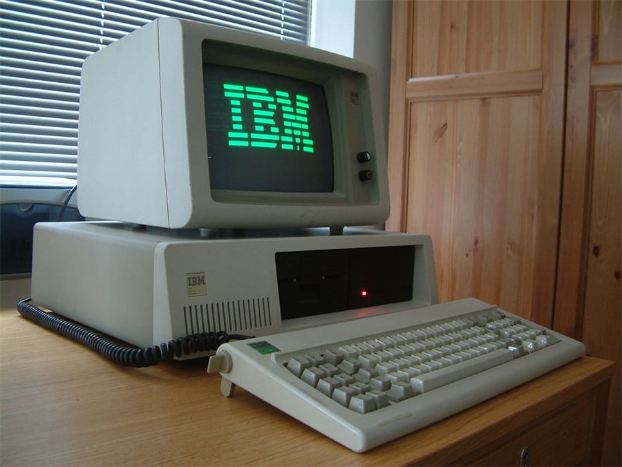 A vintage IBM personal computer with a monochrome monitor displaying “IBM,” a beige keyboard, and disk drives, sits on a wooden desk near a window and wooden cabinet.