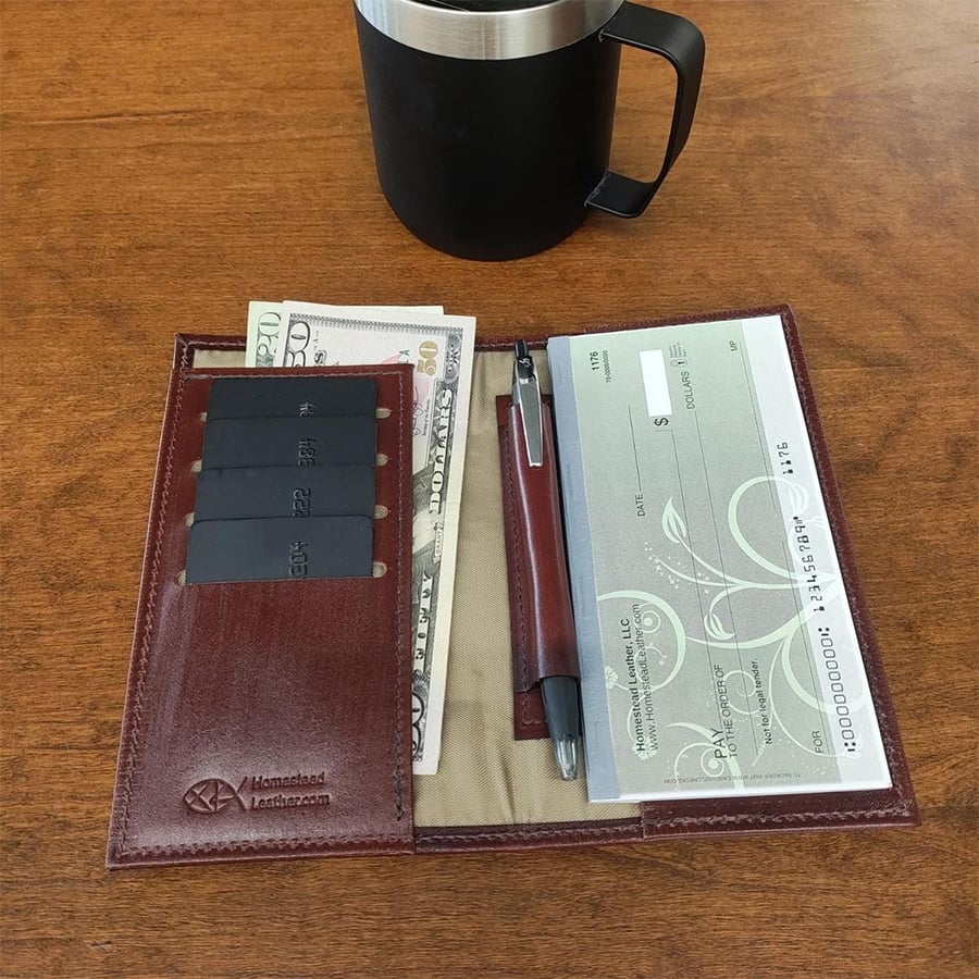 A leather checkbook holder with cards, cash, a pen, and checks sits on a wooden table. A black mug is positioned above the holder.