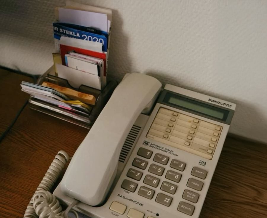 A beige landline telephone with large buttons sits on a wooden desk next to a holder filled with assorted papers, envelopes, and brochures.