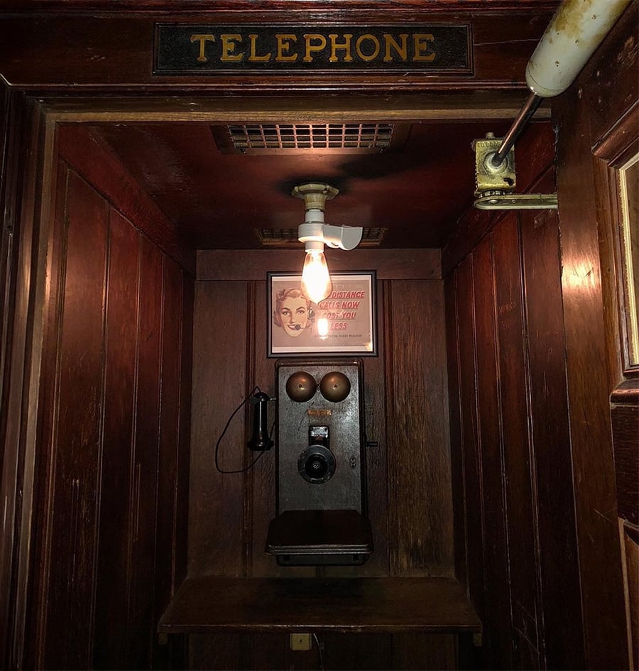 An old wooden phone booth with a rotary phone, a wooden seat, a hanging light bulb, and a “TELEPHONE” sign above the entrance. A vintage poster with a woman’s face is on the back wall.