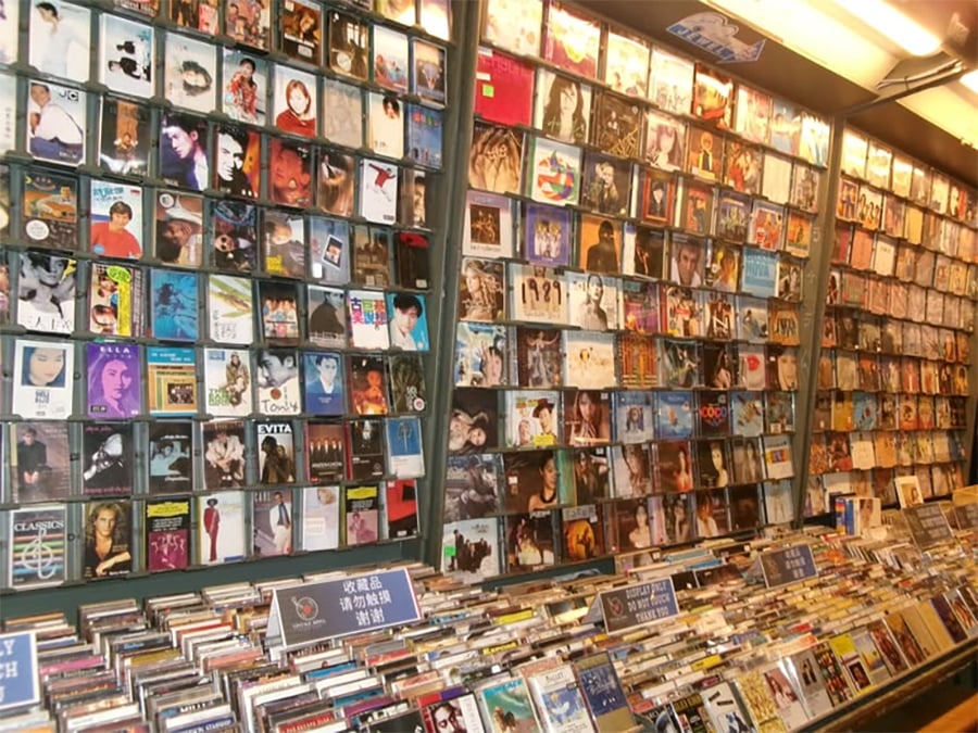 A music store display features numerous CD cases with various album covers, arranged on shelves and in bins, showcasing a wide selection of genres and artists.