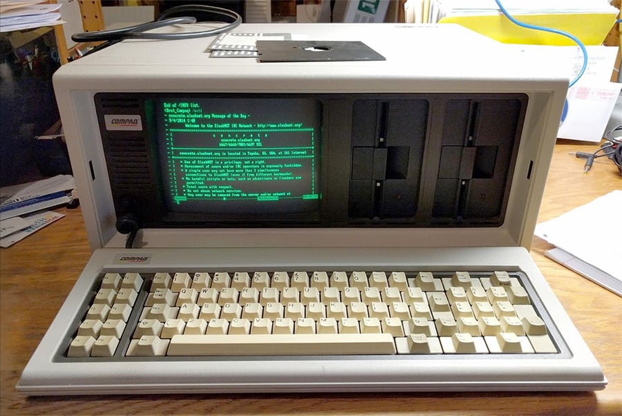 A vintage Compaq portable computer sits on a wooden desk, displaying green text on its screen, with a beige keyboard and two large floppy disk drives. Papers and other items are visible in the background.