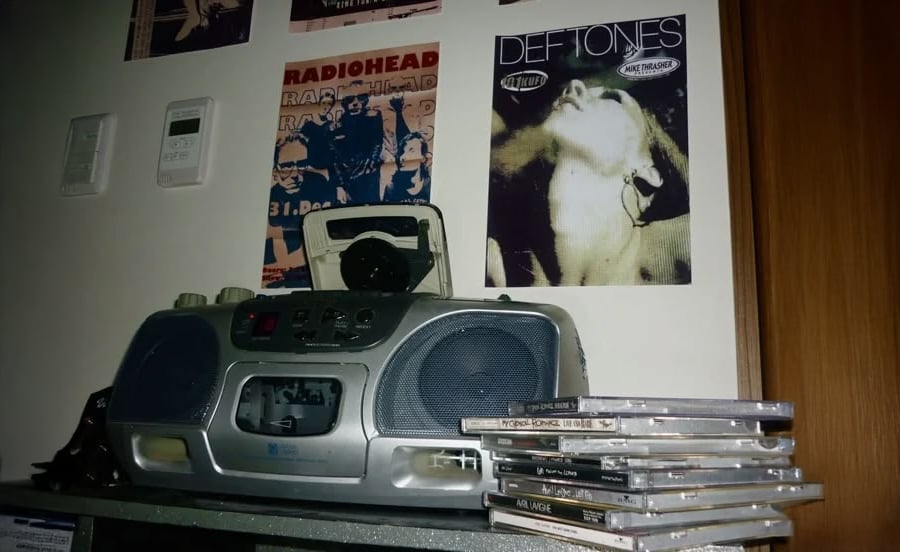 A silver boombox sits on a table with a stack of CDs beside it. On the wall above are posters, including ones for Radiohead and Deftones. The scene has a nostalgic, early 2000s vibe.