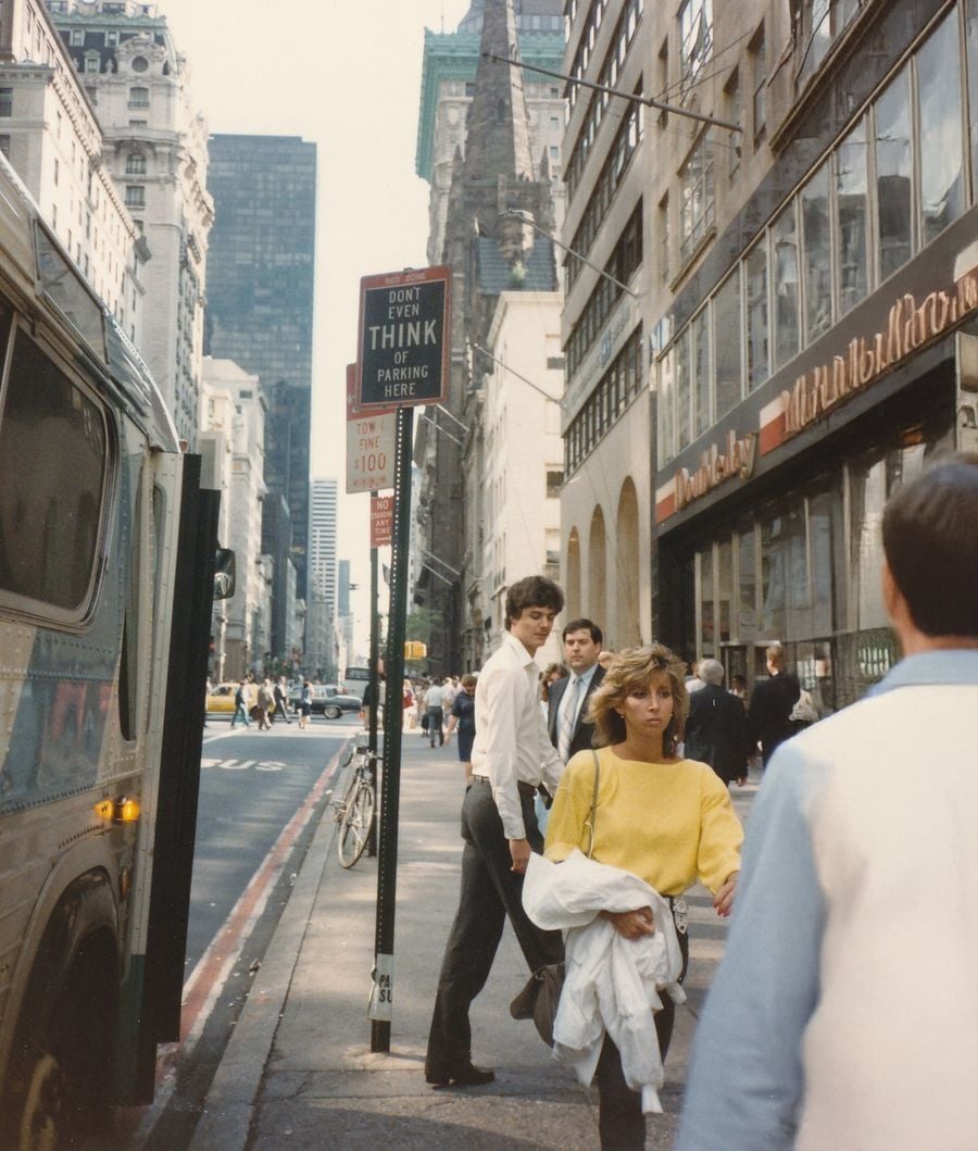 A city street scene with people walking on the sidewalk near tall buildings, a bus, and a “Don’t Even Think of Parking Here” sign. One woman in a yellow sweater looks toward the camera.