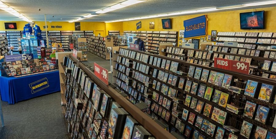 A Blockbuster video rental store interior with rows of DVDs, a blue display table, yellow walls, and signs showing categories like “KIDS” and price promotions. The store has a nostalgic, retro atmosphere.
