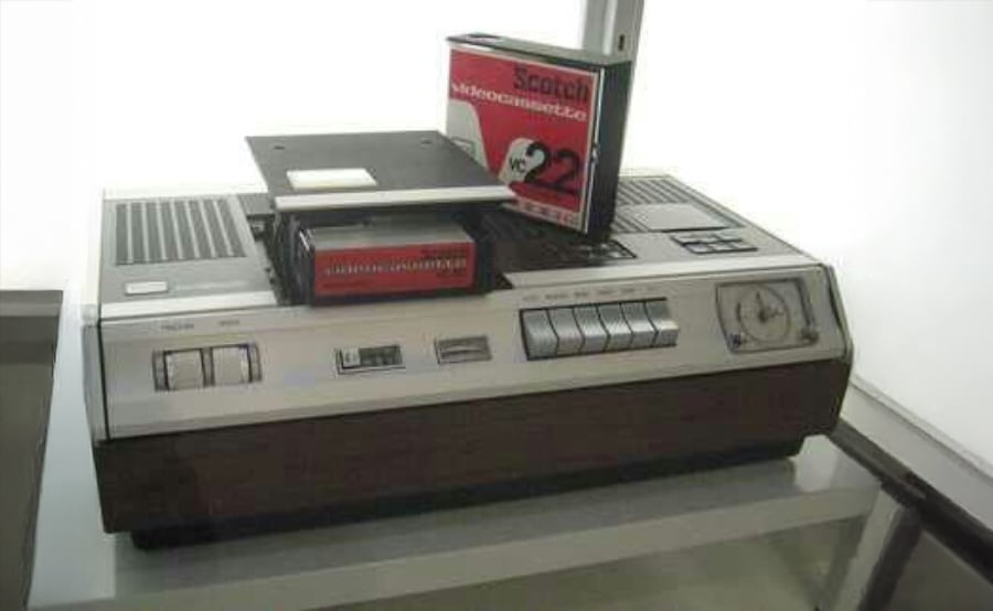 A vintage video cassette recorder sits on a table, with a large Scotch videocassette partially inserted into the front loading slot.