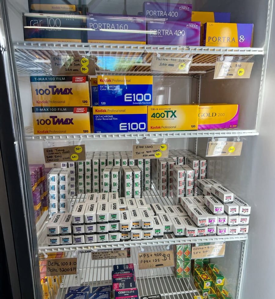A refrigerator stocked with various boxes of film rolls, including Kodak Portra, Ektachrome, Tmax, and several other brands, neatly organized on shelves with handwritten price tags.