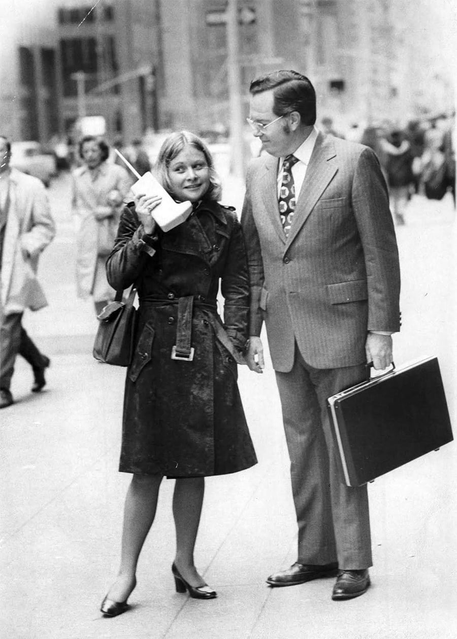 A woman in a trench coat holds a large early mobile phone to her ear, standing next to a man in a suit with a briefcase on a busy city street.