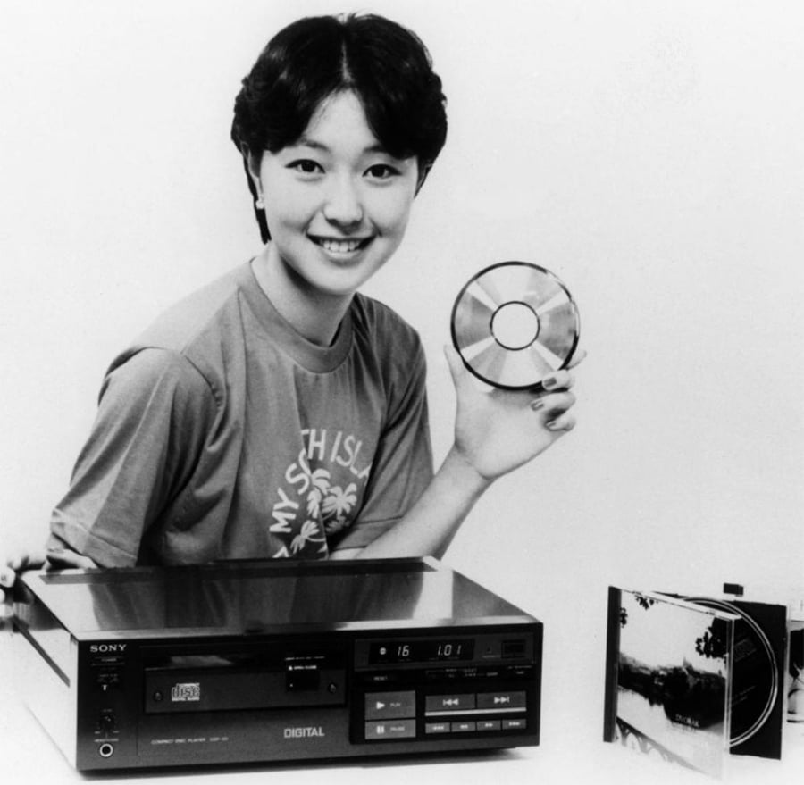 A smiling person holds up a compact disc next to a Sony CD player and an open CD case. The person wears a t-shirt and has short hair. The setting appears to be a promotional or advertising photo.