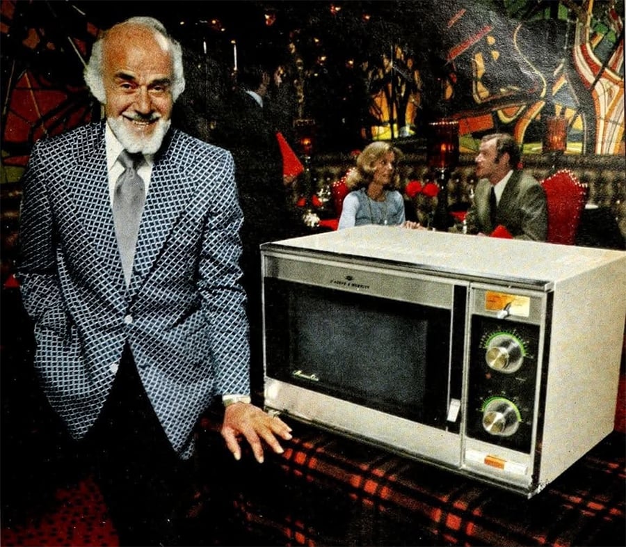 A man in a patterned suit stands beside a vintage microwave on a plaid tablecloth, with people dining and colorful abstract art in the background.