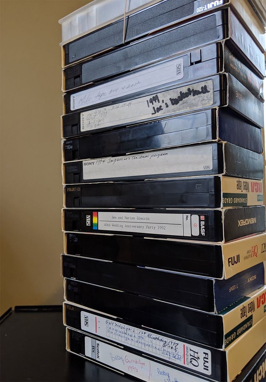 A stack of various VHS tapes, some labeled by hand with years and events like “Wedding Anniversary Party 1992,” sitting on a black surface against a beige wall.