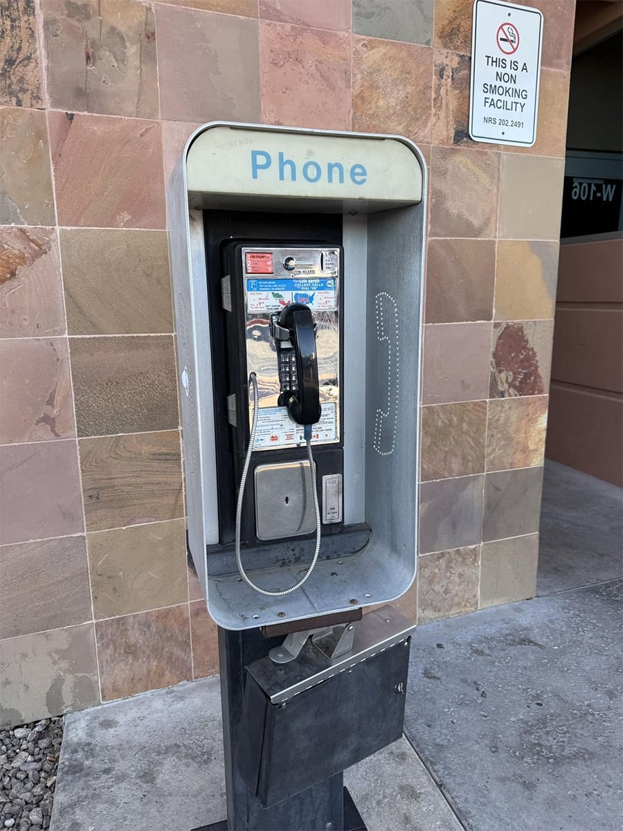 An outdoor payphone with a black handset is mounted on a pedestal against a tiled wall. A “Phone” sign is above it, and a “No Smoking” sign is visible on the wall nearby.