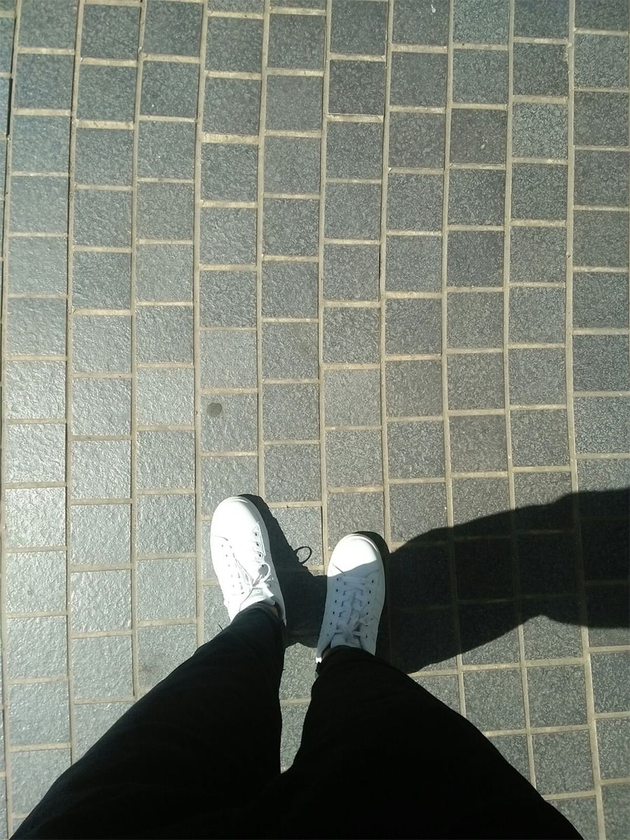 A person wearing black pants and white sneakers stands on a sunlit, tiled pavement. Their shadow is visible on the right side of the image.