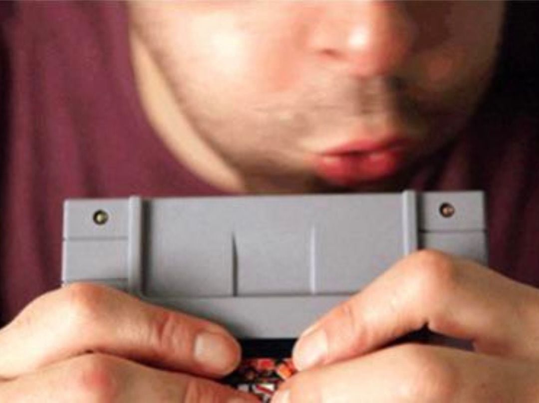 A person is blowing into a gray video game cartridge while holding it with both hands, likely attempting to fix it before use.