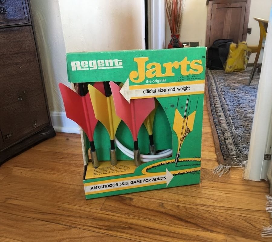 A vintage box of Regent Jarts lawn darts with red and yellow darts inside, sitting on a wooden floor in a home setting.