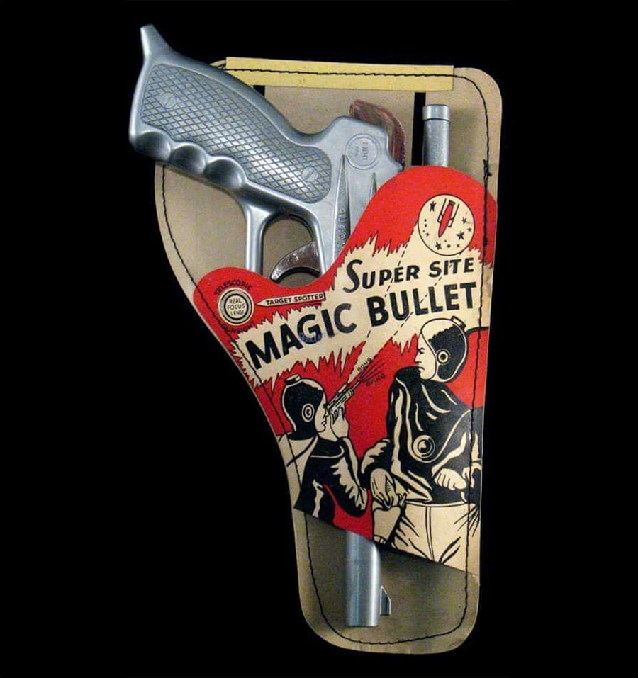 A toy silver gun in vintage packaging labeled “Super Site Magic Bullet,” showing two people in space suits, one firing the toy gun with stars and a clock in the background.