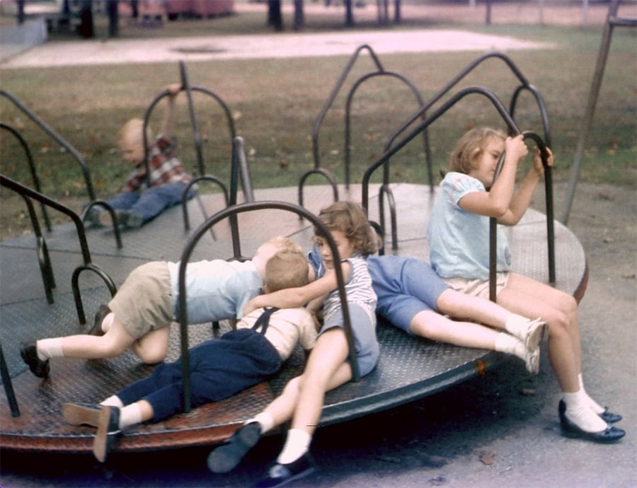 Five children lie and sit on a metal merry-go-round at a playground, holding on to the bars. The scene appears candid and playful, with trees and park equipment in the background.