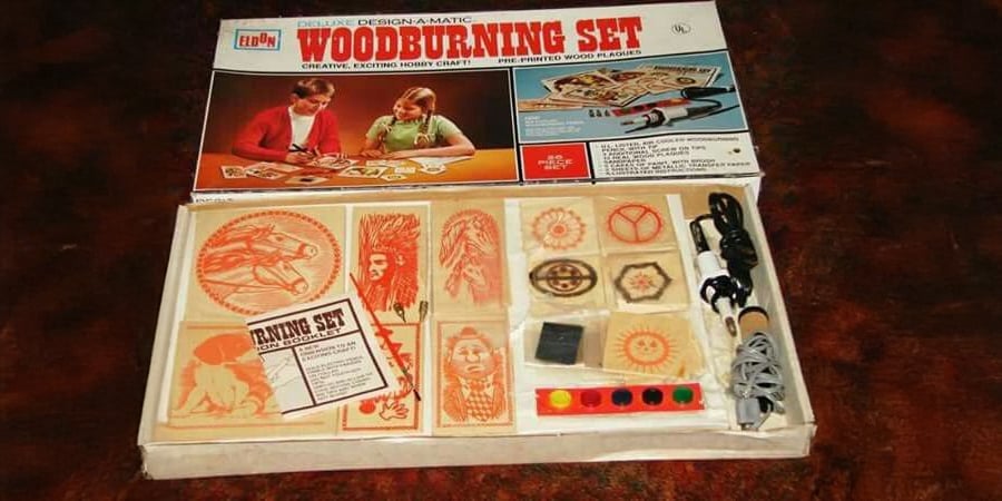 Open woodburning set box with various tools, color paints, wooden plaques featuring engraved designs, instruction booklet, and a packaging lid showing two people using the kit.