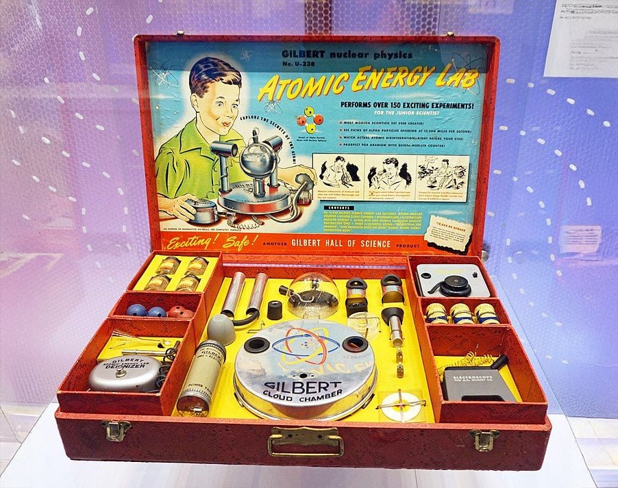 Vintage Gilbert Atomic Energy Lab science kit displayed in an open red case, showing various scientific instruments, materials, and colorful packaging with illustrations and experiment instructions.