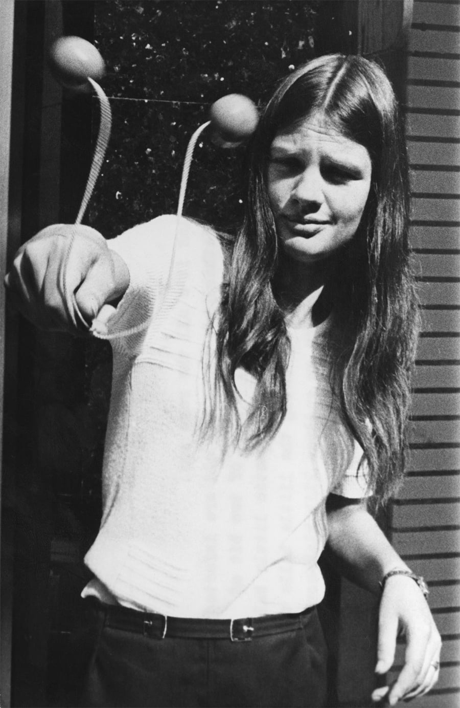 A young woman with long hair plays with two balls attached by strings to her fingers, holding them up and smiling slightly, standing outside near a wall.