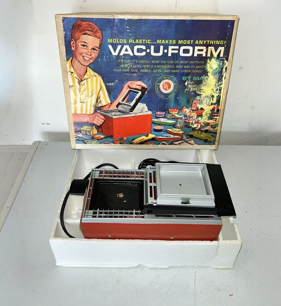 A vintage VAC-U-FORM toy set sits in its box, with colorful packaging showing a smiling boy using the machine to mold plastic shapes. The toy and accessories are neatly displayed in a white tray.