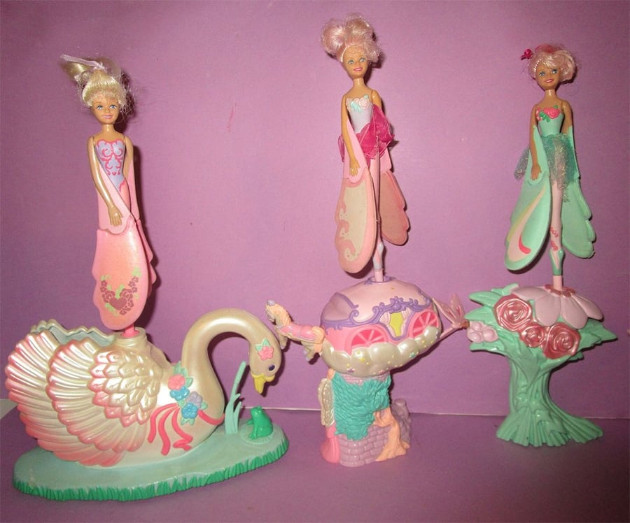 Three fairy dolls with blonde hair and colorful dresses stand on pastel bases: a swan, a spinning top, and a flower. The background is a solid light purple color.
