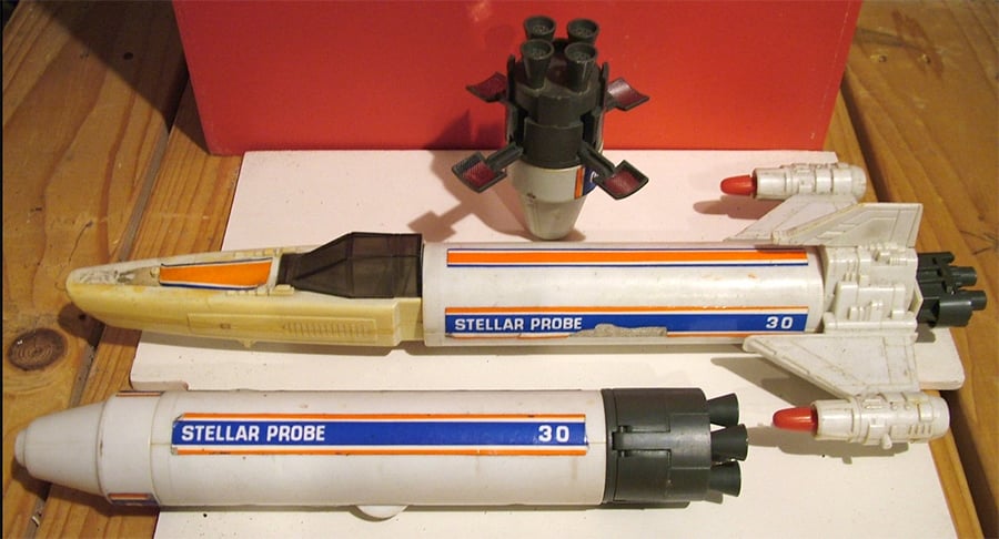Three vintage toy rockets labeled “Stellar Probe 30” are displayed on a wooden surface, with detachable parts and red-tipped details, set against a red and white background.