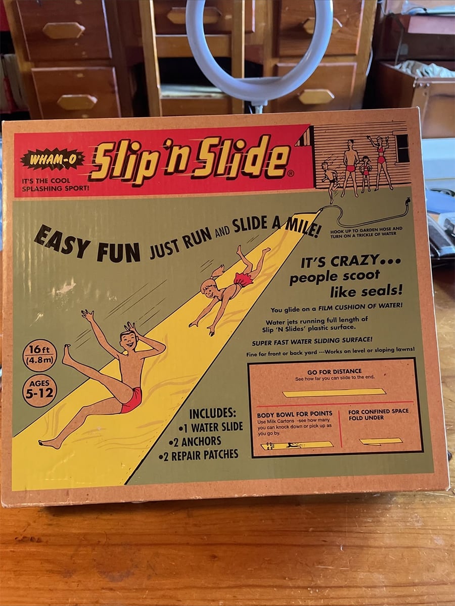 A vintage Wham-O Slip ’n Slide box features cartoon kids sliding on a yellow water slide. Text promotes easy fun for ages 5-12, includes water slide, anchors, and repair patches.