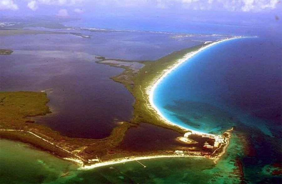 Aerial view of a narrow strip of land bordered by turquoise ocean on one side and darker inland water on the other, with some buildings at the tip.