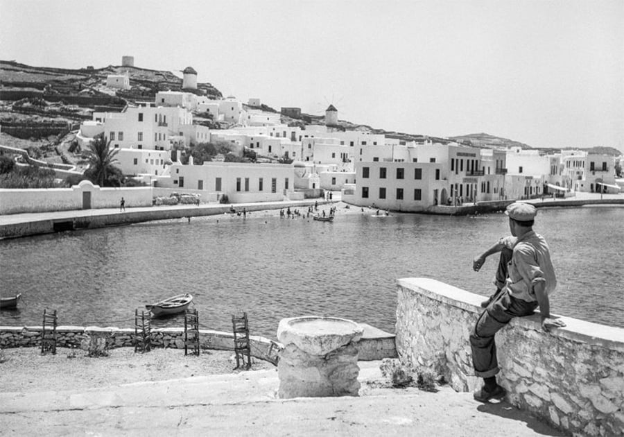 A man sits on a stone wall overlooking a calm waterfront lined with white buildings and windmills. Empty chairs and a boat are visible by the water, with people walking along the opposite shore.
