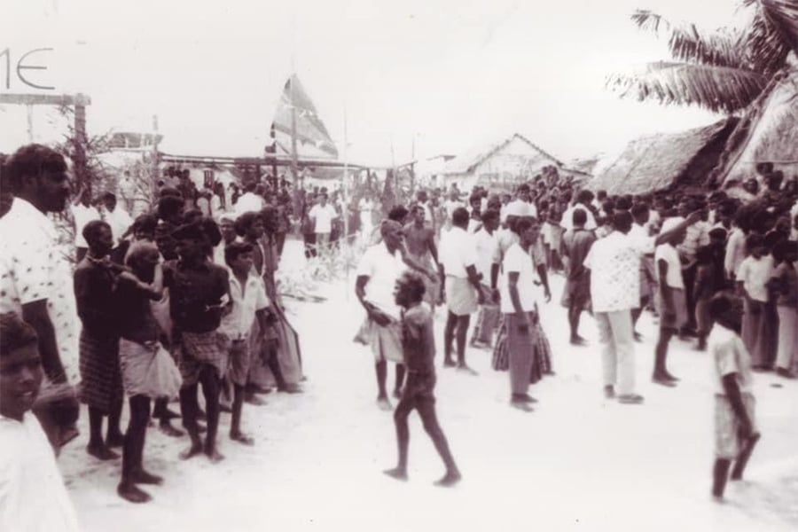 A crowd of people, including children, gather outside on a sandy area near huts and palm trees. The scene appears to be a village event or gathering. The photo is in black and white.