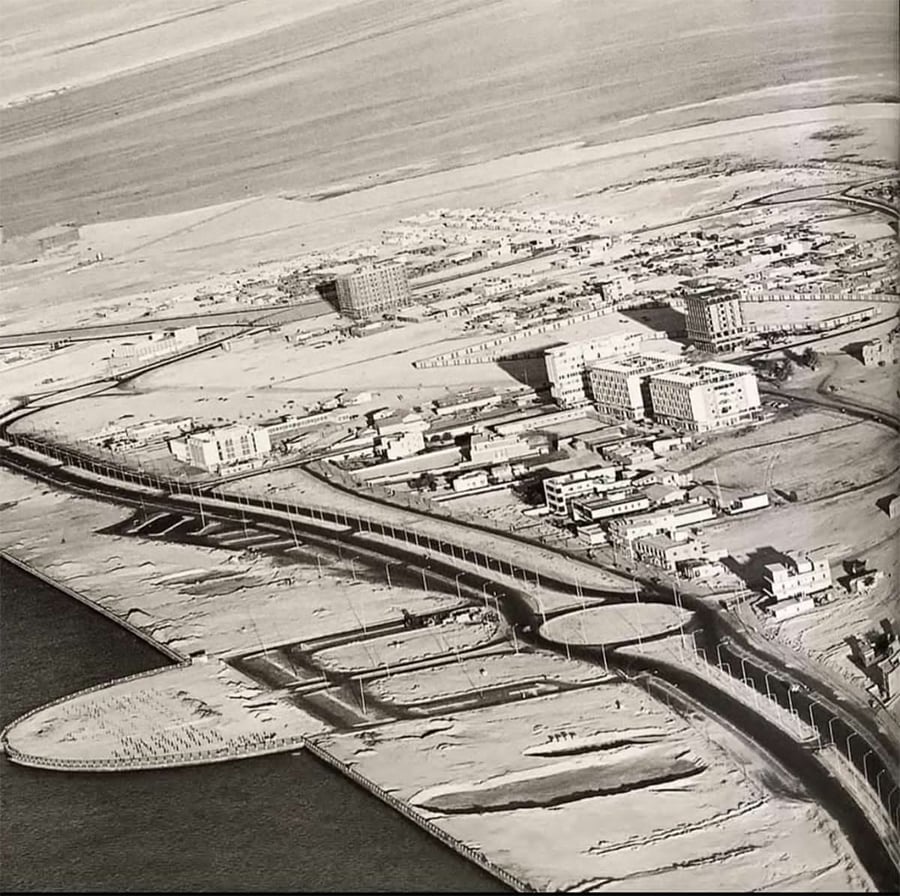 A black-and-white aerial view of a coastal city under development, with buildings, roads, and a bridge near the shoreline, surrounded by sand and water.